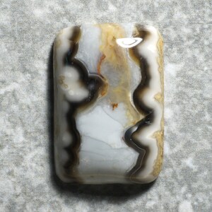 May include: A rectangular polished gemstone cabochon featuring a smooth surface with swirling patterns of opaque white, translucent gray, dark brown, and golden yellow bands that create a layered, organic appearance throughout the stone.