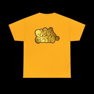 May include: Mustard yellow t-shirt with a gold-colored, bubble-style graphic that reads "SKRU SKRU". The graphic is centered on the shirt's front. The shirt has short sleeves and a crew neck.