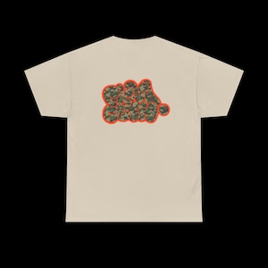 May include: A beige t-shirt with a camouflage design in the shape of the words "REAL GANG" on the back. The text is outlined in orange. The shirt has short sleeves and a crew neck.