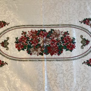 May include: A white tablecloth with a floral design. The center features an oval border with a chain-like pattern, framing a cluster of red and white poinsettias. Additional poinsettia clusters are at each corner.