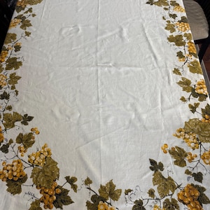 Puede incluir: Mantel blanco con un patrón repetido de hojas verdes y uvas amarillas. El diseño de uvas bordea los bordes del mantel. El mantel es rectangular y parece estar hecho de algodón o lino.