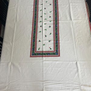 May include: A rectangular white tablecloth with a festive Christmas design. The border features a red and black stripe with holly and berry accents. The center has a rectangular design with holly and berry motifs.