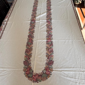 Puede incluir: Mantel rectangular color crema con un diseño central de guirnalda floral en tonos rojos, verdes y dorados. El borde presenta un patrón floral a juego. El mantel está hecho de una tela suave.