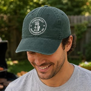 May include: A dark green baseball cap featuring a circular white graphic on the front. The design depicts a hot dog wearing a top hat with the text "CONDIMENT INDEPENDENCE DAY" printed in a circular border around the central illustration.
