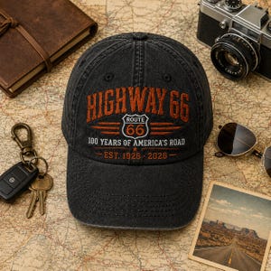 May include: A black baseball cap featuring embroidered text that reads HIGHWAY 66, ROUTE 66, 100 YEARS OF AMERICA'S ROAD, and EST. 1926 - 2026. The cap has a structured crown, a curved brim, and a distressed, washed-out fabric finish.