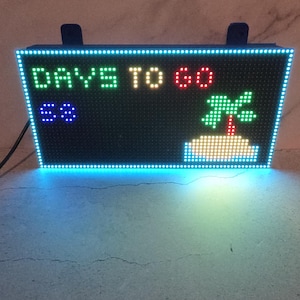 Może przedstawiać: Prostokątny wyświetlacz LED wyświetla tekst "DAYS TO GO" w kolorze zielonym, ż&oacute;łtym i czerwonym. Liczba "68" jest wyświetlana na niebiesko, a także widoczna jest zielona grafika z palmą. Wyświetlacz jest obramowany niebieską obw&oacute;dką LED.