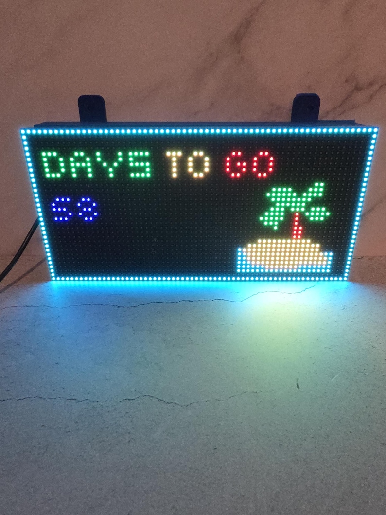 Może przedstawiać: Prostokątny wyświetlacz LED wyświetla tekst "DAYS TO GO" w kolorze zielonym, ż&oacute;łtym i czerwonym. Liczba "68" jest wyświetlana na niebiesko, a także widoczna jest zielona grafika z palmą. Wyświetlacz jest obramowany niebieską obw&oacute;dką LED.