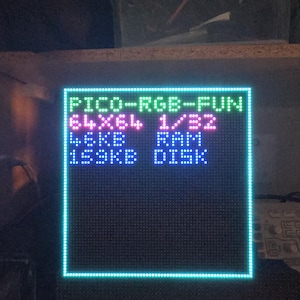 Może przedstawiać: Kwadratowy wyświetlacz LED z niebieską obw&oacute;dką i tekstem "PICO-RGB-FUN", "64X64 1/32", "4.6KB RAM" i "153KB DISK". Tekst jest w kolorze zielonym, r&oacute;żowym i białym. Wyświetlacz znajduje się na ciemnej powierzchni.