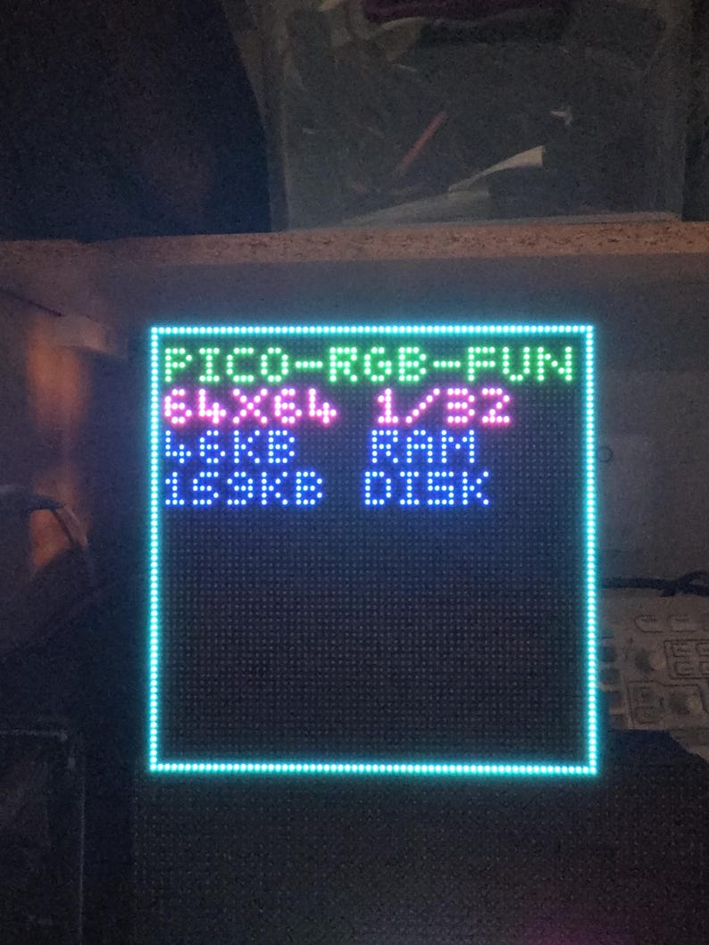 Może przedstawiać: Kwadratowy wyświetlacz LED z niebieską obw&oacute;dką i tekstem "PICO-RGB-FUN", "64X64 1/32", "4.6KB RAM" i "153KB DISK". Tekst jest w kolorze zielonym, r&oacute;żowym i białym. Wyświetlacz znajduje się na ciemnej powierzchni.