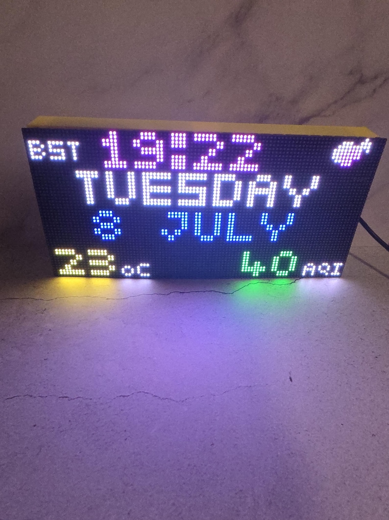 Może przedstawiać: Cyfrowy zegar wyświetlający godzinę 19:22, wtorek, lipiec, 23&deg;C i 40 AQI. Zegar ma czarną tarczę z białymi, niebieskimi, zielonymi i ż&oacute;łtymi diodami LED. G&oacute;ra i boki są ż&oacute;łte.