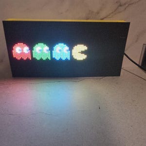 Może przedstawiać: Czarny wyświetlacz LED z pikselowymi duchami Pac-Mana w kolorze czerwonym, zielonym i niebieskim, a następnie Pac-Man w kolorze ż&oacute;łtym. Wyświetlacz ma ż&oacute;łtą g&oacute;rę i jest podłączony do źr&oacute;dła zasilania.