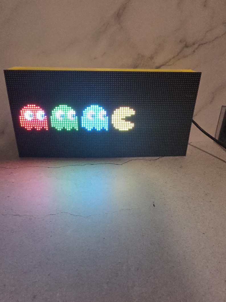 Może przedstawiać: Czarny wyświetlacz LED z pikselowymi duchami Pac-Mana w kolorze czerwonym, zielonym i niebieskim, a następnie Pac-Man w kolorze ż&oacute;łtym. Wyświetlacz ma ż&oacute;łtą g&oacute;rę i jest podłączony do źr&oacute;dła zasilania.
