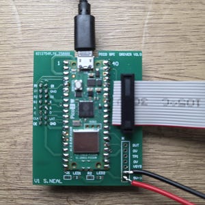 Puede incluir: Una placa de circuito verde con un cable USB negro conectado. La placa tiene varios componentes electrónicos, incluidos chips, conectores y pines etiquetados. También se adjuntan un cable de cinta y cables rojo y negro.