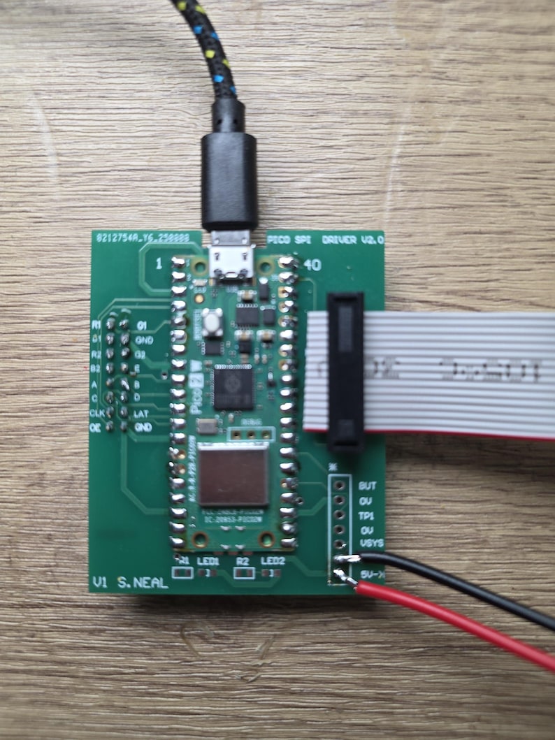 Może przedstawiać: Zielona płytka drukowana z podłączonym czarnym kablem USB. Płytka posiada r&oacute;żne elementy elektroniczne, w tym układy scalone, złącza i oznaczone piny. Dołączony jest r&oacute;wnież kabel taśmowy oraz czerwone i czarne przewody.