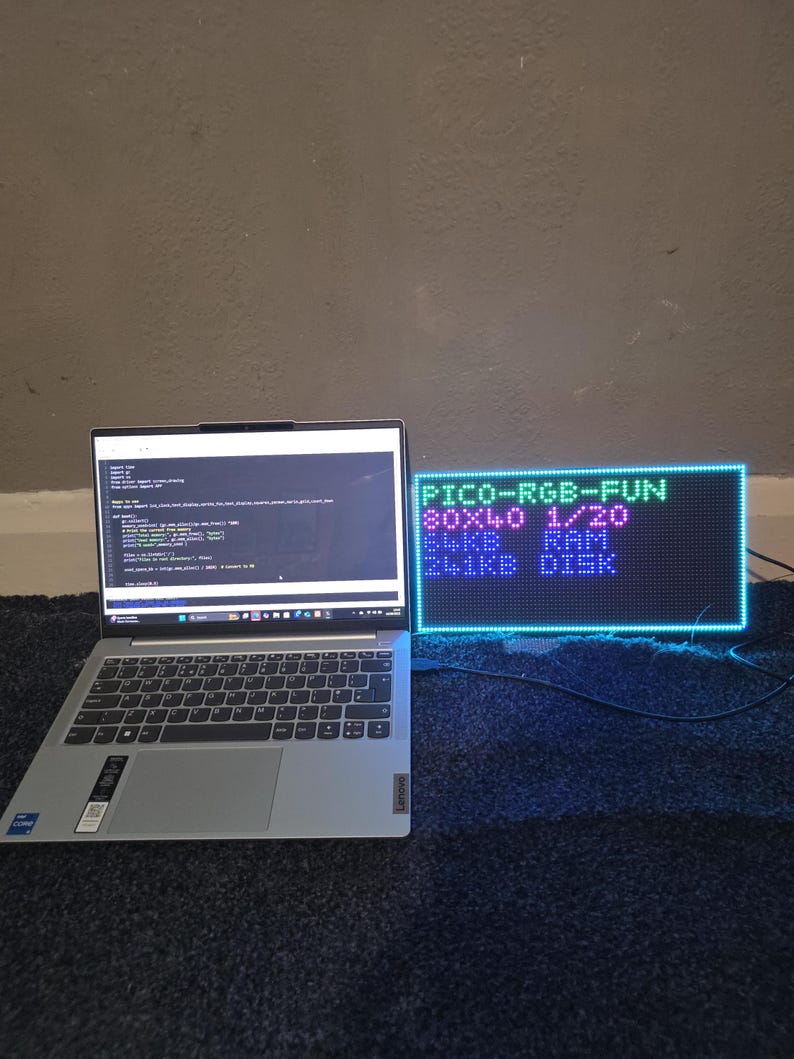 Może przedstawiać: Srebrny laptop z czarną klawiaturą stoi na ciemnoniebieskiej powierzchni obok prostokątnego wyświetlacza LED. Na wyświetlaczu widnieje napis "PICO-RGB-FUN" i inne specyfikacje techniczne w r&oacute;żnych kolorach. Ekran laptopa wyświetla linie kodu.