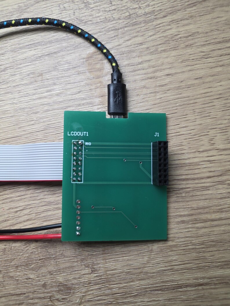 Może przedstawiać: Zielona płytka drukowana z nadrukiem "LCDOUT1" i "J1". Czarny kabel USB jest podłączony do płytki. Podłączona jest r&oacute;wnież taśma i czerwone i czarne przewody.