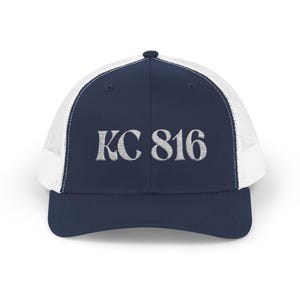 May include: A navy blue trucker cap with a white mesh back. The front of the cap features the embroidered text "KC 816" in silver. The cap has a curved peak and a button on top.