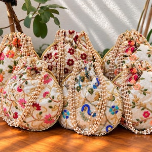 Puede incluir: Una colección de seis bolsas con cordón de color blanco roto que presentan un intrincado bordado floral en tonos vibrantes de rosa, rojo, naranja, azul y verde. Cada bolsa está decorada con un asa hecha de múltiples hilos de perlas blancas.