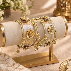 May include: A set of gold-toned bangles featuring clear acrylic bands adorned with metallic gold floral accents and small dangling beads. The bracelets are displayed on a cylindrical beige velvet stand with gold metal end caps and a rectangular base.