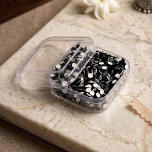 May include: A clear, square plastic container filled with numerous small, round, black and white craft eyes. The container features a removable, transparent lid that is partially lifted, revealing the collection of tiny, circular components inside.