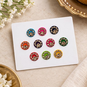 May include: A set of ten circular adhesive bindi stickers arranged on a white card. Each sticker features a vibrant felt base in colors including blue, pink, orange, purple, green, and yellow, decorated with a ring of small clear rhinestones.