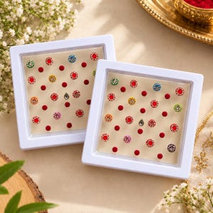 May include: Two square white display cases containing an assortment of adhesive face stickers. The collection features small circular red dots alongside decorative gems in various colors including green, blue, yellow, and pink, arranged in a grid.