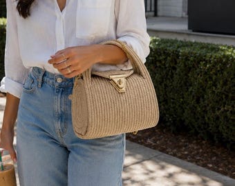 May include: A beige cylindrical handbag crafted from coiled rope material, featuring a gold-toned metal turn-lock closure on the front flap and a matching rope handle. The bag has a structured, rounded shape with vertical texture throughout the exterior.