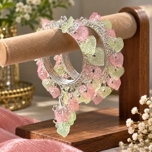 May include: Two silver-toned bangles featuring a textured metallic band adorned with dangling translucent leaf-shaped charms in shades of pale pink and light green. The charms are attached to the circular frame with small silver-colored metal rings.