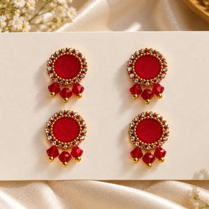 May include: Two pairs of circular stud earrings featuring a vibrant red center disc surrounded by a border of small clear crystals. Each earring is accented with three dangling red faceted beads suspended from the bottom edge.