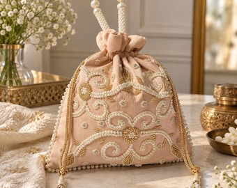May include: A light peach-colored drawstring pouch bag featuring intricate gold thread embroidery and white pearl bead embellishments. The bag includes a top handle made of white pearls and two long gold-toned braided drawstrings with pearl tassels.