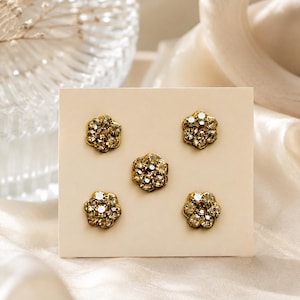 May include: A set of five flower-shaped stud earrings or buttons arranged on a rectangular display card. Each piece features a gold-toned metal base set with multiple clear, faceted, sparkling crystals arranged in a circular floral pattern.