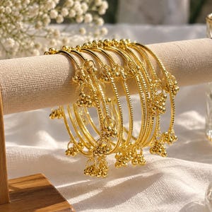 May include: A set of gold-toned metal bangles featuring a textured, ridged surface. Each bangle is adorned with multiple small, dangling bell-shaped charms attached to circular rings, creating a layered, metallic appearance on a display stand.