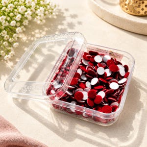 May include: A clear, square plastic container with a partially open lid, filled with numerous small, circular, deep red velvet-textured adhesive dots. Each dot features a vibrant red fabric top and a plain white adhesive backing.