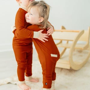 May include: Two young children wearing matching rust-colored ribbed pajamas with cream-colored cuffs and ankle cuffs. They are embracing each other. A wooden rocking toy is in the background.