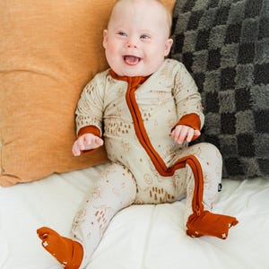 Könnte beinhalten: Ein lächelndes Baby in einem beigefarbenen und rostfarbenen Strampler mit Fuß und einem Muster zum Thema Natur. Der Strampler hat einen durchgehenden rostfarbenen Reißverschluss und passende Bündchen und Füße. Das Baby sitzt auf einem weißen Bett mit orangefarbenen und grauen Kissen.