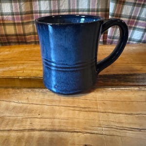 May include: A handmade, dark blue ceramic mug with a curved handle. The mug has a glossy finish and two horizontal grooves near the top. It sits on a wooden surface, with a plaid fabric in the background.