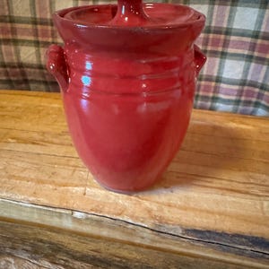 May include: A red ceramic jar with a lid. The jar has a rounded body with two small handles and a lid with a pointed finial. The jar is sitting on a wooden surface.