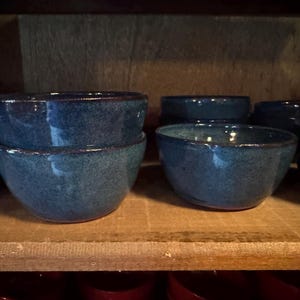 May include: A collection of small, round ceramic bowls with a speckled blue glaze and dark brown rims. The bowls feature a smooth, curved shape and are arranged in a group, with some stacked on top of each other, displaying a consistent finish.