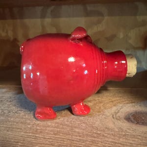 May include: A glossy red ceramic pig-shaped bottle stopper with a cork. The pig has small legs, a tail, and a snout. The stopper is on a wooden surface.