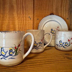 May include: Three off-white ceramic mugs and one matching small plate, each featuring hand-painted blue and yellow birds perched on blue wavy lines with red and green cattail accents. The ceramic pieces have a rustic, textured finish.