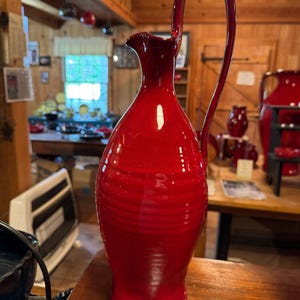 May include: A tall, red ceramic pitcher with a curved handle and a pouring spout. The pitcher has a glossy finish and a ribbed texture. The background shows a rustic interior with other red ceramic items.