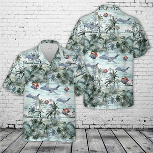 May include: A short-sleeve button-down shirt featuring a light blue and green tropical palm tree pattern interspersed with gray military transport aircraft and small red circular emblems throughout the fabric design on both the front and back.