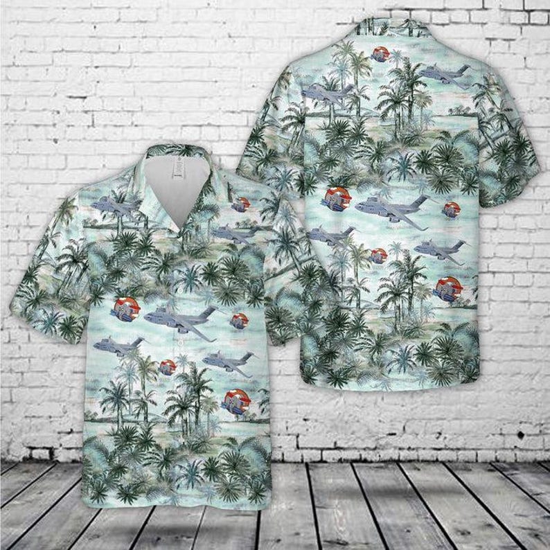 May include: A short-sleeve button-down shirt featuring a light blue and green tropical palm tree pattern interspersed with gray military transport aircraft and small red circular emblems throughout the fabric design on both the front and back.