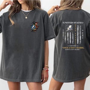 May include: Dark gray t-shirt with a distressed American flag and Navy Seabee emblem on the front. The back features a flag design with text: "UNITED STATES NAVY SEABEE DUTY." The shirt also has the text: "ONCE A NAVY SEABEE, ALWAYS A NAVY SEABEE."