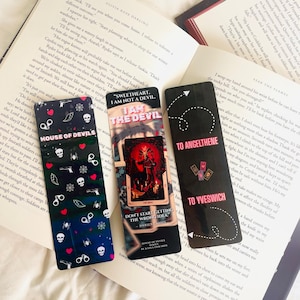 Könnte beinhalten: Drei dekorative Lesezeichen mit dunklem Hintergrund und weißem Text auf einem offenen Buch. Die Lesezeichen zeigen verschiedene Designs, darunter Totenköpfe, Herzen und den Text "SWEETHEART, I AM NOT A DEVIL."
