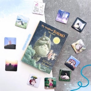 Może przedstawiać: Kolekcja kwadratowych magnesów z scenami i postaciami z "Mój sąsiad Totoro" oraz egzemplarz powieści. Magnesy prezentują różne ilustracje Totoro i innych postaci w żywych kolorach. Okładka książki przedstawia tytuł i dużą ilustrację Totoro.