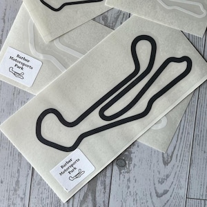 May include: Several white stickers with black outlines of a race track are displayed. The stickers have the text "Barber Motorsports Park" and a small track graphic. The stickers are on a light gray wooden surface.