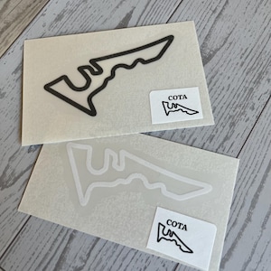May include: Two stickers featuring the outline of a racetrack in black and white. Each sticker has a small rectangular label with the word "COTA" and a smaller racetrack outline. The stickers are on a light-colored background.
