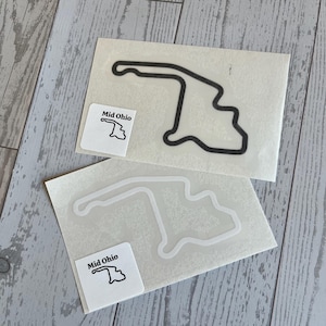 May include: Two stickers featuring the outline of the Mid-Ohio Sports Car Course racetrack. One sticker has a black outline, and the other has a white outline. Each sticker has a small square label with the text "Mid Ohio" and a track outline.
