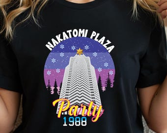 May include: Black t-shirt featuring a graphic of a skyscraper with the text "Nakatomi Plaza Party 1988". The design includes a purple and blue gradient background with snowflakes and trees. The shirt is a crew neck.